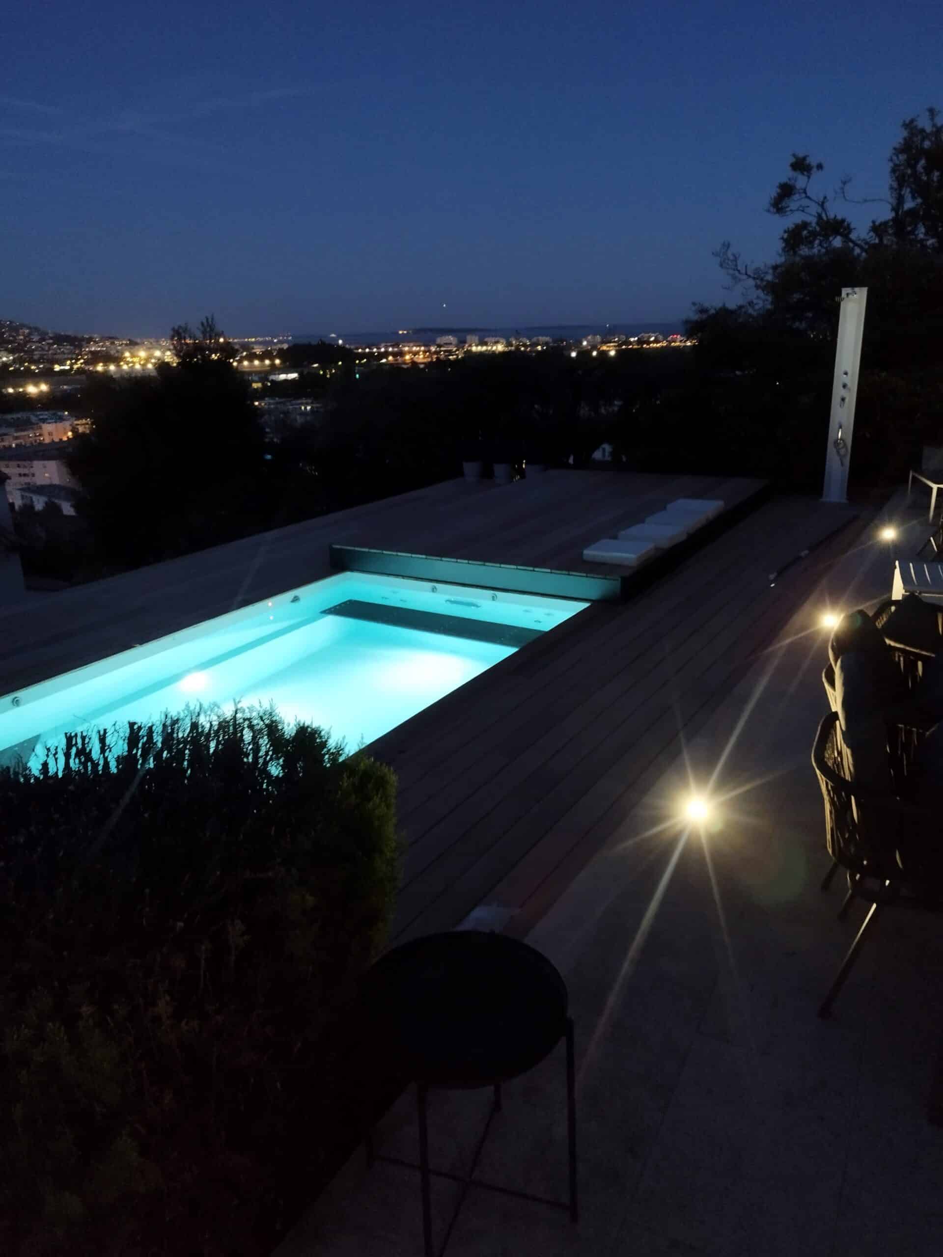 Piscine de Luxe avec Vue sur Ville la Nuit | Terrasse Bois Piscine éclairée sur une terrasse en bois éclaire a nuit avec vue panoramique sur les lumières de la ville.